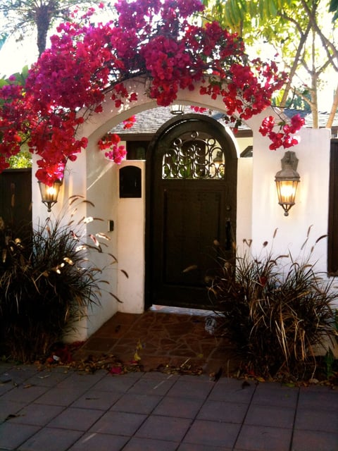 Arched entryway with black door framed by vibrant magenta bougainvillea flowers, white walls, decorative lanterns, and ornamental grasses