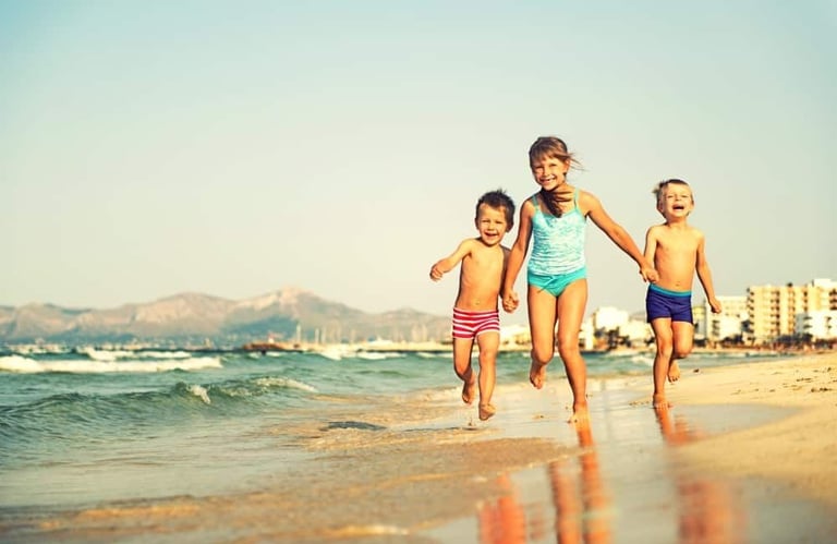 Three young children running together on a sunny beach with mountains and buildings visible in the background