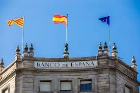 Ornate Bank of Spain building facade with Spanish, regional, and institutional flags flying from the roof against blue sky